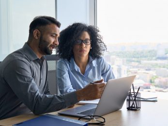 Multiethnic colleagues sitting at desk looking at laptop computer in office. Commercial solutions for patient engagement and HCP communications
